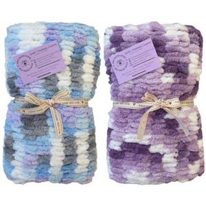 Puede incluir: Dos mantas de bebé de crochet hechas a mano, una en tonos de azul, gris y blanco, y la otra en tonos de morado, blanco y lavanda. Ambas mantas están atadas con una cinta marrón y una etiqueta que dice "Sarah's Handmade Creations".