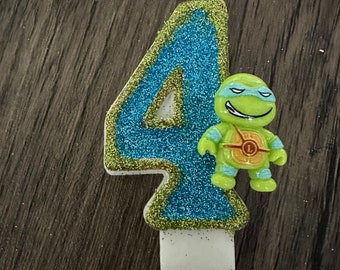May include: A number 4 birthday candle with a blue glitter design and a green and yellow trim. A small Teenage Mutant Ninja Turtle figure is attached to the candle. The candle is white at the base.