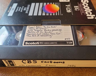Könnte beinhalten: Ein schwarzes VHS-Band mit einem Etikett, auf dem "Scotch EG VIDEOCASSETTE" und "CBS cartoons with commercials 2 hours 1986" steht. Das Band ist mit einer handgeschriebenen Liste von Shows beschriftet, darunter "Muppet Babies "By the Book"", "Noisy Neighbors", "Hulk Hogans Rockin Wrestling", "Battle of the Bands", "Amazons Just wanna have fun" und "The Art of Wrestling".