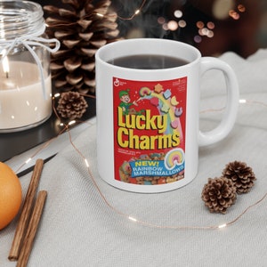 May include: White ceramic mug featuring a Lucky Charms cereal box design. The design includes the words "Lucky Charms" in yellow, a leprechaun, and colorful marshmallow shapes. The mug is filled with a dark liquid.