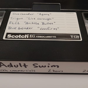 Puede incluir: Una cinta VHS negra etiquetada como "Adult Swim" con el texto "with commercials 2 hours 2003". La cinta está etiquetada con el título "Scotch EG VIDEOCASSETTE T-120" y enumera los siguientes programas: "Blue Gender "Agony"", "Trigun "Live through"", "FLCL "Brittle Bullet"", y "Blue Gender "confirm"".