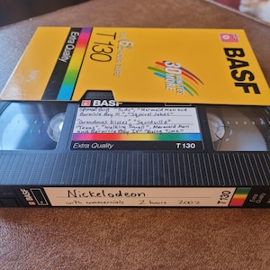 Puede incluir: Una cinta VHS amarilla con el texto "BASF Extra Quality T130 2 HOUR TAPE" y "30 MINUTES MORE". La cinta está etiquetada "Nickelodeon with commercials 2 hours 2002" y enumera episodios del programa "SpongeBob SquarePants".