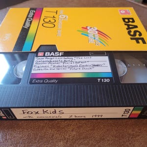 以下が含まれることがあります： 「The 6 Hour Tape」と「30 Minutes More」の文字が印刷された黄色のVHSテープ。テープには「BASF」と「T130」のラベルが貼られています。テープには「Fox Kids」というタイトルと「with commercials 2 hours 1999」というテキストが書かれています。