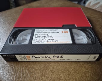 Pode incluir: Uma videocassete VHS Fuji T-120 preta com uma parte superior vermelha. O rótulo diz "Barney PBS" e lista títulos de músicas. A fita está rotulada com "2 horas 30 min" e o ano de "1992".