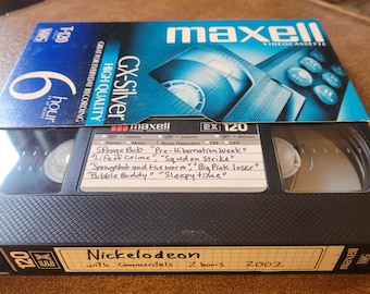 Puede incluir: Una cinta VHS negra con una etiqueta azul que dice "Maxell Videocassette" y "EX 120". La cinta está etiquetada con "Nickelodeon with Commercials 2 hours 2002" y una lista manuscrita de episodios de "Sponge Bob": "Pre-Hibernation Week", "Life of Crime", "Squid on Strike", "Spongebab and the worm", "Big Pink loser", "Bubble Buddy", "Sleepy time".