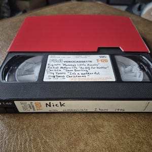 May include: A black Fuji T-120 VHS videotape with a red top. The label includes handwritten titles like "Rugrats" and "Tiny Toons Christmas." The tape is labeled "Nick" and "1996."