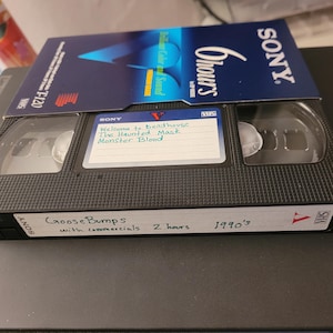 以下が含まれることがあります： 白いラベルに「Goosebumps with commercials 2 hours 1990's」と書かれた黒いVHSテープ。テープには「Welcome to Deadhouse The Haunted Mask Monster Blood」というタイトルが付けられています。