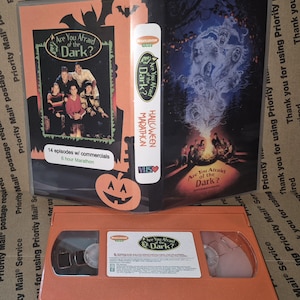 Peut inclure: Une cassette VHS orange et son boîtier assorti pour le Are You Afraid of the Dark? Halloween Marathon. Le boîtier présente un groupe de personnes, un feu de camp et une illustration de fantôme. La cassette a une étiquette blanche avec du texte noir.