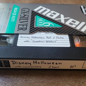 May include: A VHS tape labeled "Disney Halloween" with "with commercials 2 hours 80's" written on the label. The tape is a Maxell GX-Silver T-120 VHS tape.