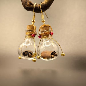 Könnte beinhalten: Ein Paar einzigartige Ohrringe mit kleinen, durchsichtigen Glasflaschen mit Korken. In jeder Flasche befindet sich eine aufgerollte Schriftrolle. Die Flaschen sind mit goldenem Draht, roten Perlen und goldfarbenen Akzenten verziert und hängen an goldfarbenen Ohrhaken.