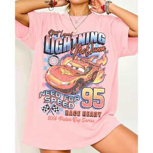 May include: Pink t-shirt featuring a graphic of Lightning McQueen from the movie Cars. The design includes the text "Street Legend LIGHTNING McQueen", "Need for Speed", "95", and "Race Ready". The shirt is from the 2006 Piston Cup Series.