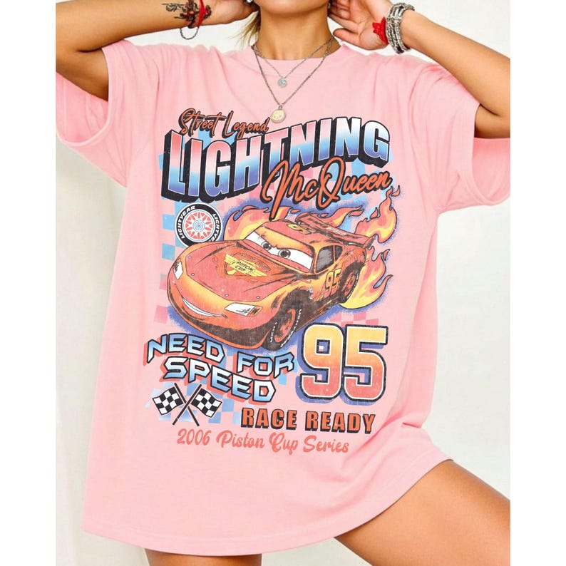 May include: Pink t-shirt featuring a graphic of Lightning McQueen from the movie Cars. The design includes the text "Street Legend LIGHTNING McQueen", "Need for Speed", "95", and "Race Ready". The shirt is from the 2006 Piston Cup Series.
