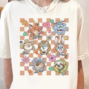 Puede incluir: Camiseta color crema con un gr&aacute;fico colorido de personajes de Zootopia, cada uno rodeado de dise&ntilde;os florales. Los nombres "Nick Wilde", "Judy Hopps" y otros est&aacute;n impresos debajo de los personajes sobre un fondo a cuadros marr&oacute;n y blanco.
