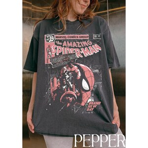 Puede incluir: Camiseta gris oscuro con una portada de c&oacute;mic vintage de Spider-Man. El gr&aacute;fico incluye el texto "AMAZING SPIDER-MAN" y "MARVEL COMICS GROUP". La camiseta tiene un aspecto desgastado y un cuello redondo cl&aacute;sico.