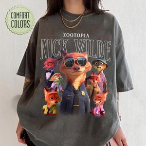 Puede incluir: Camiseta gris oscuro con un gr&aacute;fico de Nick Wilde de Zootopia con gafas de sol, rodeado de otros personajes. Los textos "ZOOTOPIA" y "NICK WILDE" est&aacute;n impresos encima del gr&aacute;fico. La camiseta tiene un logotipo de "COMFORT COLORS".