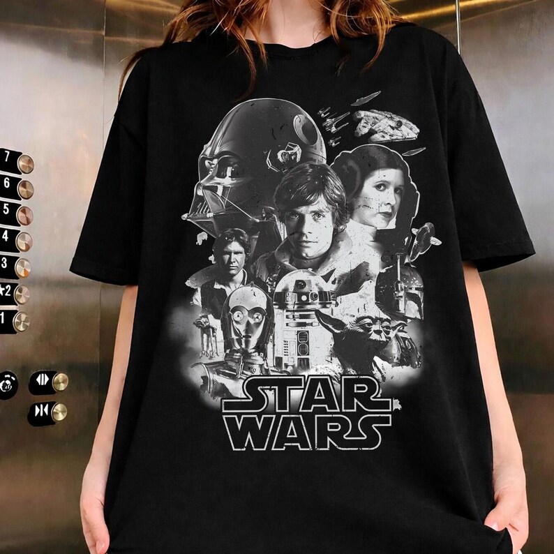 Op de afbeelding: Zwart T-shirt met een Star Wars-afbeelding. Het ontwerp bevat Darth Vader, Luke Skywalker, prinses Leia, Han Solo, C-3PO, R2-D2, Yoda en het Star Wars-logo in wit. Het shirt heeft een ronde hals.