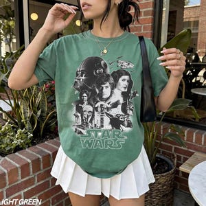 Op de afbeelding: Lichtgroen Star Wars grafisch t-shirt met Darth Vader, Luke Skywalker, prinses Leia en andere personages. Het shirt heeft een ronde hals en korte mouwen. De tekst "STAR WARS" is in het wit gedrukt.