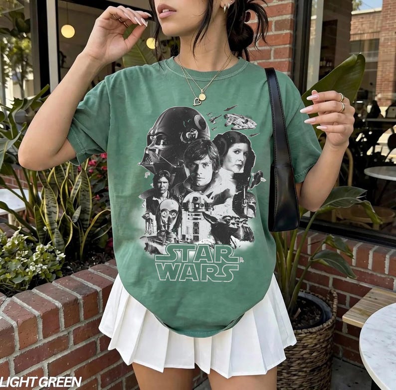 Op de afbeelding: Lichtgroen Star Wars grafisch t-shirt met Darth Vader, Luke Skywalker, prinses Leia en andere personages. Het shirt heeft een ronde hals en korte mouwen. De tekst "STAR WARS" is in het wit gedrukt.