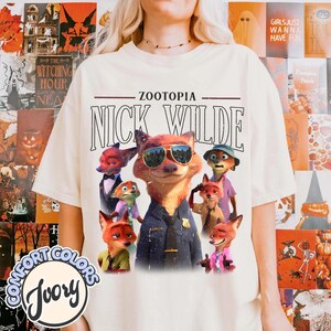 Puede incluir: Camiseta de color marfil con un gr&aacute;fico de Nick Wilde de Zootopia con gafas de sol, rodeado de otros personajes. El texto "ZOOTOPIA" y "NICK WILDE" est&aacute;n encima del gr&aacute;fico. La camiseta tiene el logo de Comfort Colors.