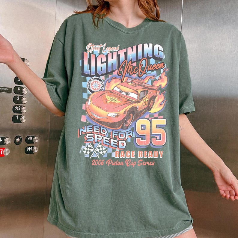 May include: Green t-shirt featuring a graphic of Lightning McQueen from the movie Cars. The design includes the text "Lightning McQueen", "Need for Speed", and the number "95". The shirt is a casual, oversized style.