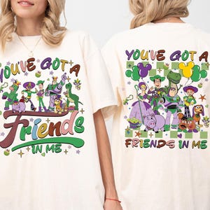 May include: Cream-colored t-shirt featuring a colorful graphic of Toy Story characters with the text "YOU'VE GOT A FRIENDS IN ME". The design includes Buzz Lightyear, Woody, and other characters. The back of the shirt also has a similar design.