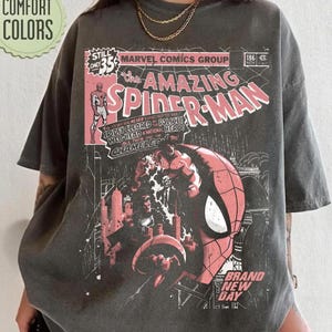 Puede incluir: Camiseta gris oscuro con una portada de c&oacute;mic de The Amazing Spider-Man. El dise&ntilde;o incluye el personaje de Spider-Man, texto de c&oacute;mic y las palabras "Brand New Day". La camiseta tambi&eacute;n tiene las palabras "Comfort Colors" en un c&iacute;rculo verde.