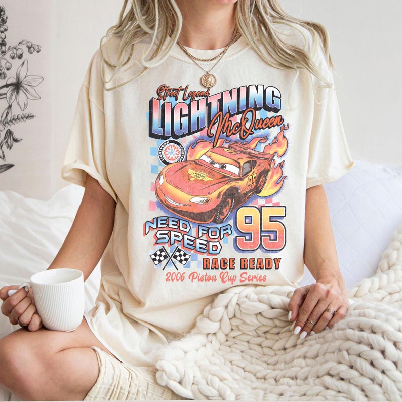 May include: Cream-colored t-shirt featuring a graphic of Lightning McQueen from the movie Cars. The design includes the text "Lightning McQueen", "Need for Speed", and "95". The shirt also has a checkered flag design and "2006 Piston Cup Series" text.