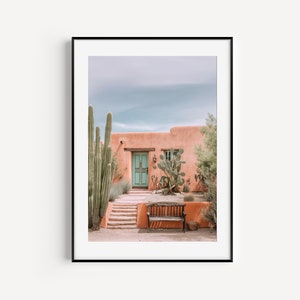 May include: A pink adobe house with a teal door and a wooden bench in front of it. There are tall cacti and other desert plants growing around the house.