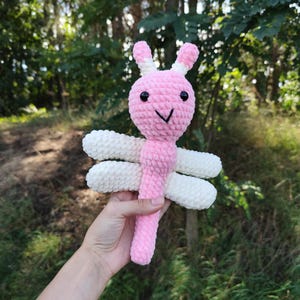 Op de afbeelding: Een roze en wit gehaakt libellespeelgoed. De libelle heeft een roze lichaam, kop en staart, met witte vleugels. De kop heeft zwarte ogen en een gestikte glimlach. Het speelgoed wordt vastgehouden tegen een achtergrond van groen gebladerte.