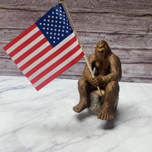 Può includere: Una statuina marrone di Bigfoot seduta su un ceppo d'albero, che tiene una piccola bandiera americana. La bandiera presenta strisce rosse e bianche e un campo blu con stelle bianche. La statuina ha un'espressione sorridente.