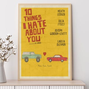 Puede incluir: Un póster enmarcado de la película "10 Things I Hate About You" con fondo amarillo. El póster presenta un SUV verde y un coche rojo, un corazón y los nombres de los actores. El texto "I burn, I pine, I perish" está en la parte inferior.
