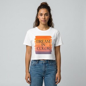 Peut inclure: Haut court blanc avec le texte "DREAM IN COLOR" en lettres noires sur un fond orange, violet et jaune de style aquarelle. Le haut est associé à un jean bleu.