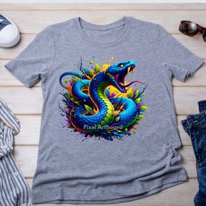 Puede incluir: Una camiseta gris jaspeado con un gráfico vibrante de una serpiente azul y verde enroscada con la boca abierta, rodeada de salpicaduras de pintura colorida y formas abstractas. El texto Pixel Artboard es visible en el centro del diseño.