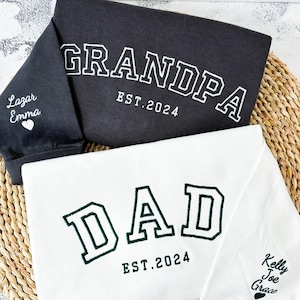 Peut inclure: Deux chemises à manches longues, une noire et une blanche, avec du texte brodé. La chemise noire dit "GRANDPA EST. 2024" et la chemise blanche dit "DAD EST. 2024". Les deux chemises ont de petits cœurs brodés sur les manches.