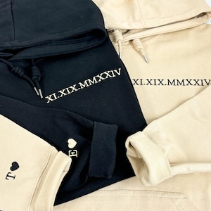 Könnte beinhalten: Zwei Kapuzenpullover, einer schwarz und einer beige, mit dem Text "XI.XIX.MMXXIV" auf der Vorderseite bestickt. Der schwarze Pullover hat einen beigefarbenen Ärmel mit einem Herz und dem Buchstaben "E" bestickt. Der beigefarbene Pullover hat einen schwarzen Ärmel mit einem Herz und dem Buchstaben "T" bestickt.