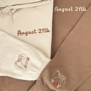 Könnte beinhalten: Zwei beigefarbene Hoodies mit gesticktem Text. Ein Hoodie sagt "August 21th" und hat ein pinkfarbenes Herz mit zwei Händen, die sich am kleinen Finger versprechen, auf dem Ärmel. Der andere Hoodie sagt "August 21th" und hat zwei Hände, die sich am kleinen Finger versprechen, mit einem roten Herzen auf dem Ärmel.