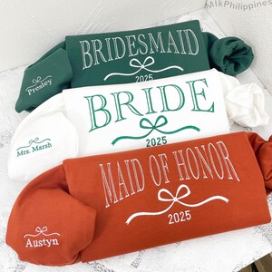May include: Three sweatshirts in green, white, and orange, each with wedding-related text. The green sweatshirt says "Bridesmaid" with "2025" below. The white sweatshirt says "Bride" with "2025". The orange sweatshirt says "Maid of Honor" with "2025".