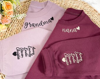 Op de afbeelding: Twee sweatshirts, één bordeauxrood en één lichtpaars, met geborduurde harten en namen. Het bordeauxrode sweatshirt zegt "nana" en het lichtpaarse sweatshirt zegt "grandma". Beide sweatshirts hebben een lijst met namen geborduurd op de mouw.