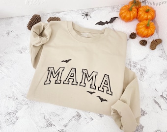 May include: Beige long-sleeved sweatshirt with the word "MAMA" in black, outlined with a spiderweb design. Three black bat silhouettes are below the word. The sweatshirt is decorated with small pumpkins and pinecones.