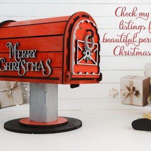 May include: A red wooden mailbox with the words "Merry Christmas" in silver lettering. The mailbox has a decorative design and sits on a gray post and black base. A reindeer figurine is to the right.