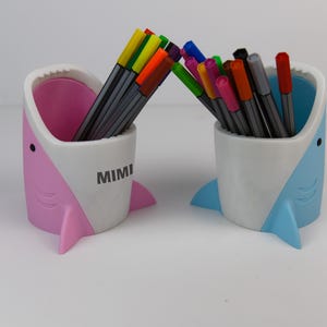 May include: Two shark-shaped pencil holders, one pink and one blue, with colorful markers inside. The pink holder has the word "MIMI" printed on the side.