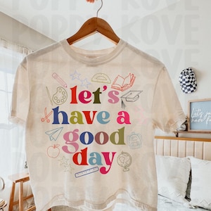 May include: A beige t-shirt with a colorful graphic design that says "Let's have a good day". The graphic includes school supplies like a pencil, ruler, paint palette, scissors, a book, a graduation cap, a paper airplane, an apple, a backpack, and a globe.