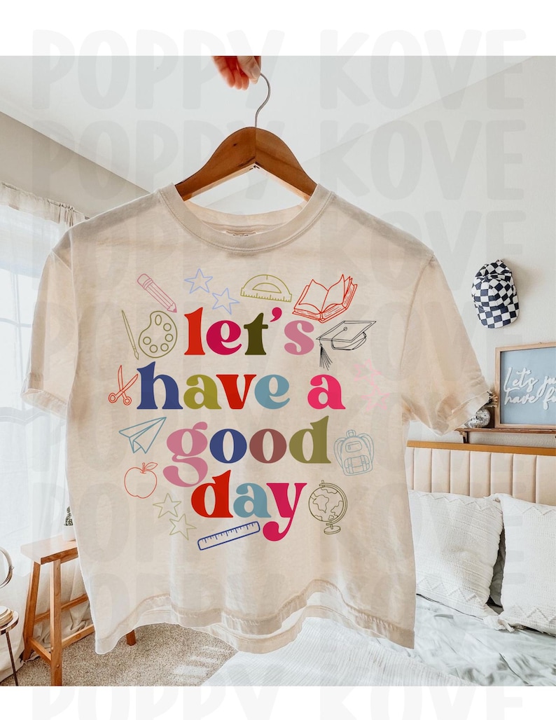 May include: A beige t-shirt with a colorful graphic design that says "Let's have a good day". The graphic includes school supplies like a pencil, ruler, paint palette, scissors, a book, a graduation cap, a paper airplane, an apple, a backpack, and a globe.