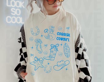 May include: White t-shirt with a blue graphic design of a mermaid, seashells, starfish, cowboy boots, a horseshoe, a hat, and a surfboard. The text "coastal cowgirl" is printed on the shirt.