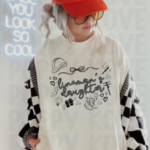 Könnte beinhalten: Weißes Langarmshirt mit dem Text "lineman & daughter" und zugehörigen Grafiken. Das Shirt zeigt einen Helm, Stiefel und Elektrowerkzeuge. Die Person trägt eine rote Kappe und eine Brille.