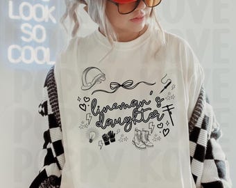 May include: White long-sleeve shirt with the text "lineman & daughter" and related graphics. The shirt features a hard hat, boots, and electrical tools. The person is wearing a red cap and glasses.