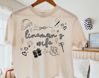 May include: A cream-colored t-shirt with a black graphic design that says "lineman's wife" and includes various symbols related to the profession, such as a hard hat, boots, and tools.