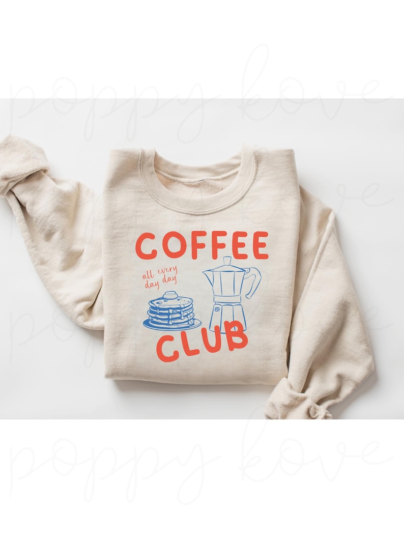 May include: A beige sweatshirt with the text "COFFEE all every day day! CLUB" in red and blue. The graphic includes a blue illustration of a coffee pot and a stack of pancakes.