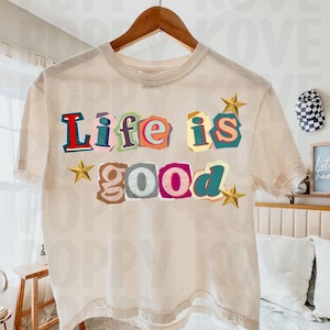May include: A beige t-shirt with the words "Life is good" in colorful, cut-out style letters. Gold star accents are placed around the text. The shirt is hanging on a wooden hanger.