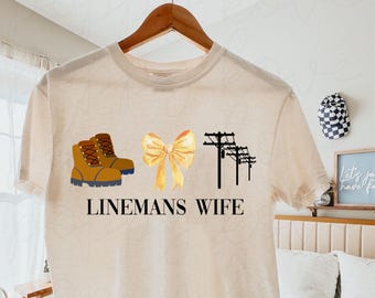 May include: A beige t-shirt with a graphic design of brown work boots, a yellow bow, and power lines. The text "Linemans Wife" is printed below the graphic.